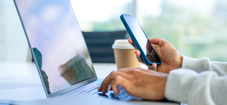 A person on a laptop with a mobile in their hand and a cup of coffee Decorative image. A person on a laptop with a mobile in their hand and a cup of coffee