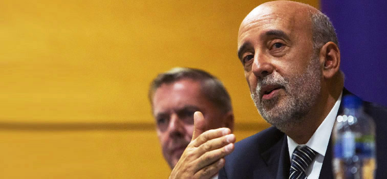 Governor Gabriel Makhlouf wearing a suit and tie pictured speaking with one hand raised with one person to his right. A yellow and blue wall is behind him.